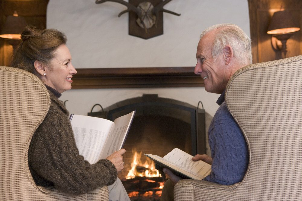 A man and a woman sit by a fireplace reading a book.